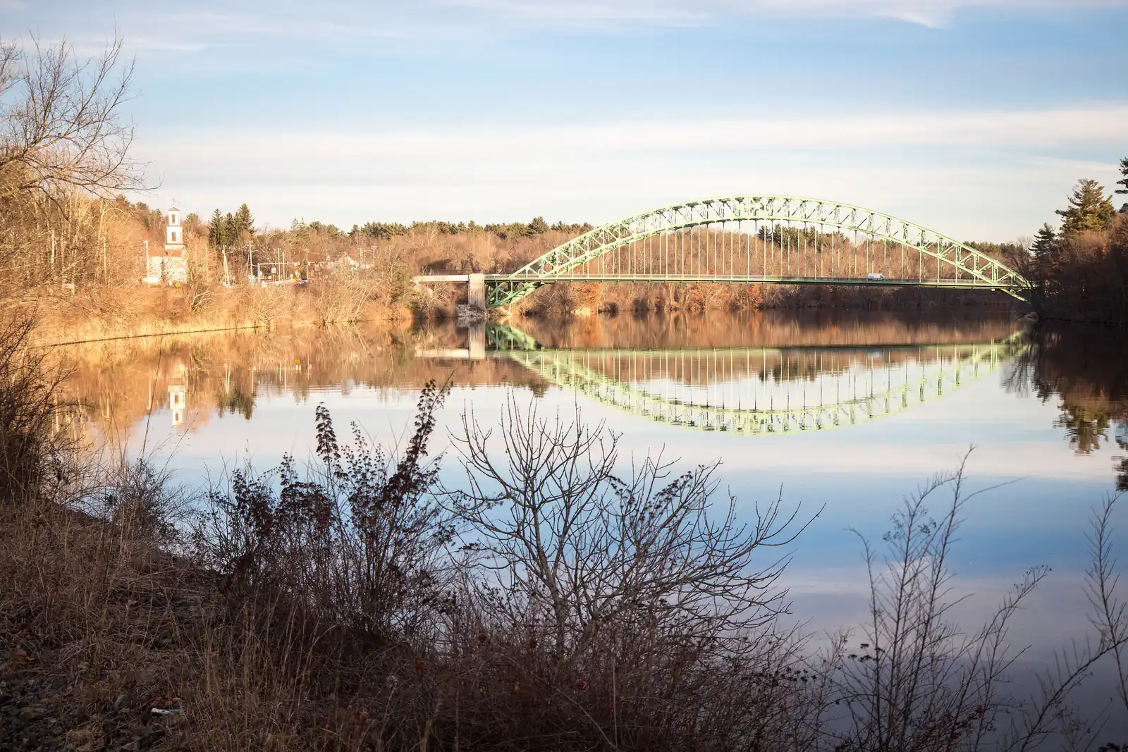 The green arch of the Tyngsborough Bridge reflected in the calm Merrimack River at golden hour